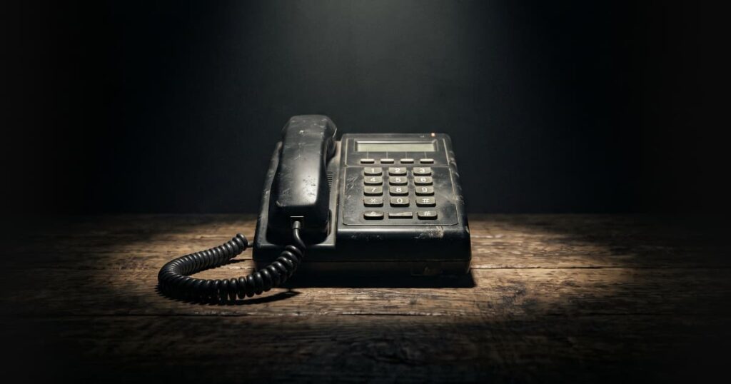 Telefone corporativo antigo e pesado iluminado sobre uma mesa vazia e escura, representando o retorno imediato ao trabalho braçal de ligar para clientes antigos.